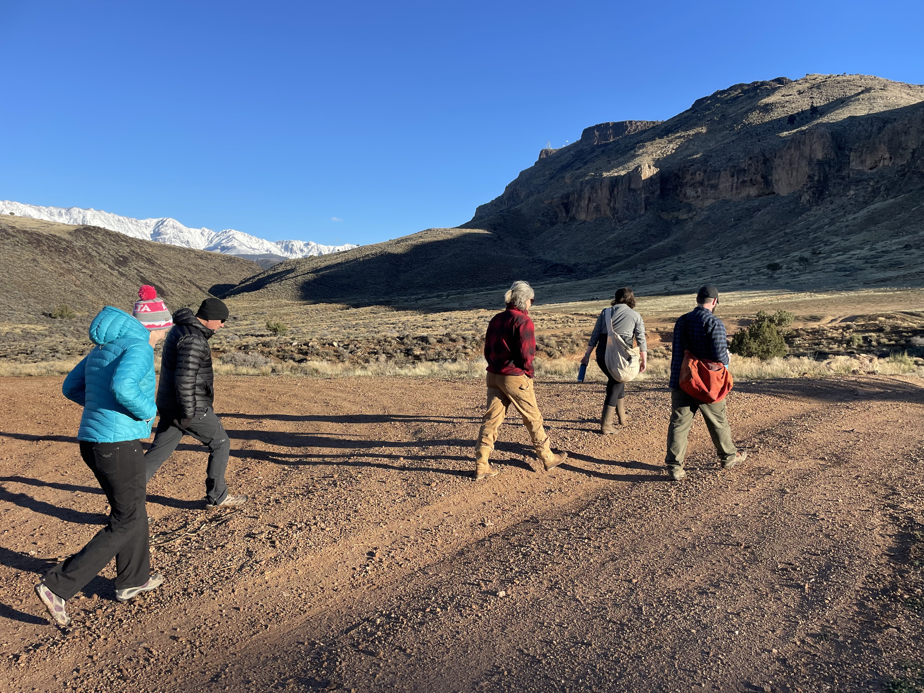 Five people hike across a dusty lot surrounded by mesas and mountains.