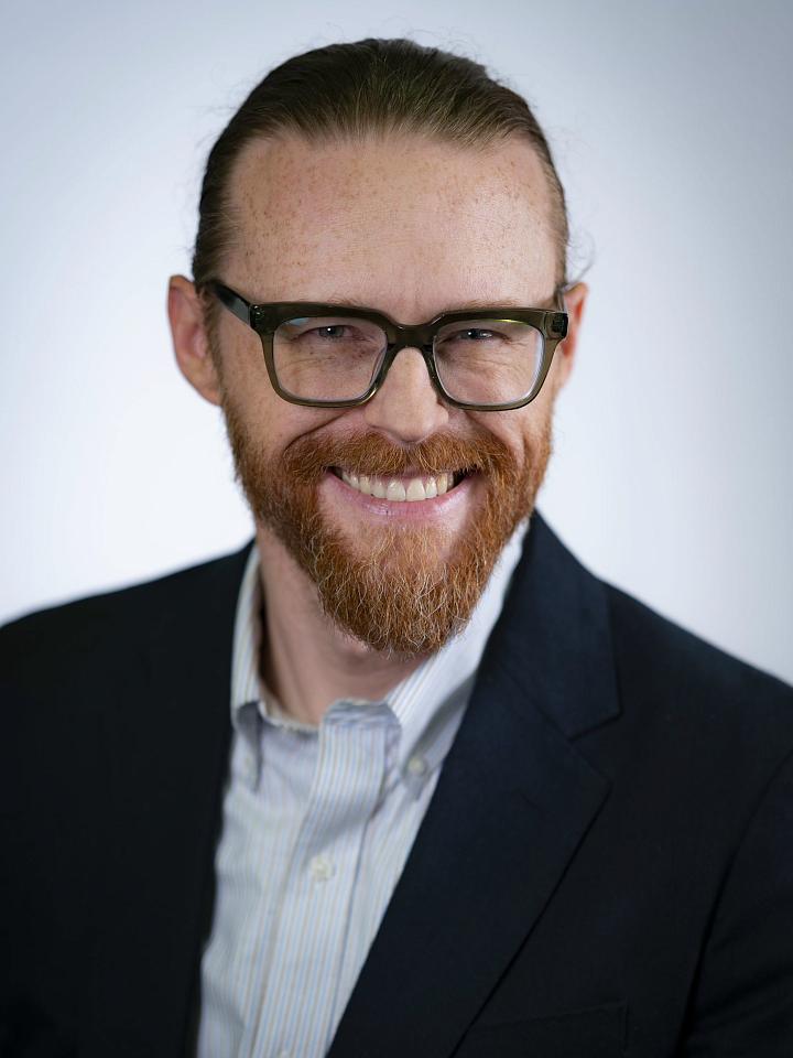 Person with glasses, freckled skin, and a red beard smiles at the camera