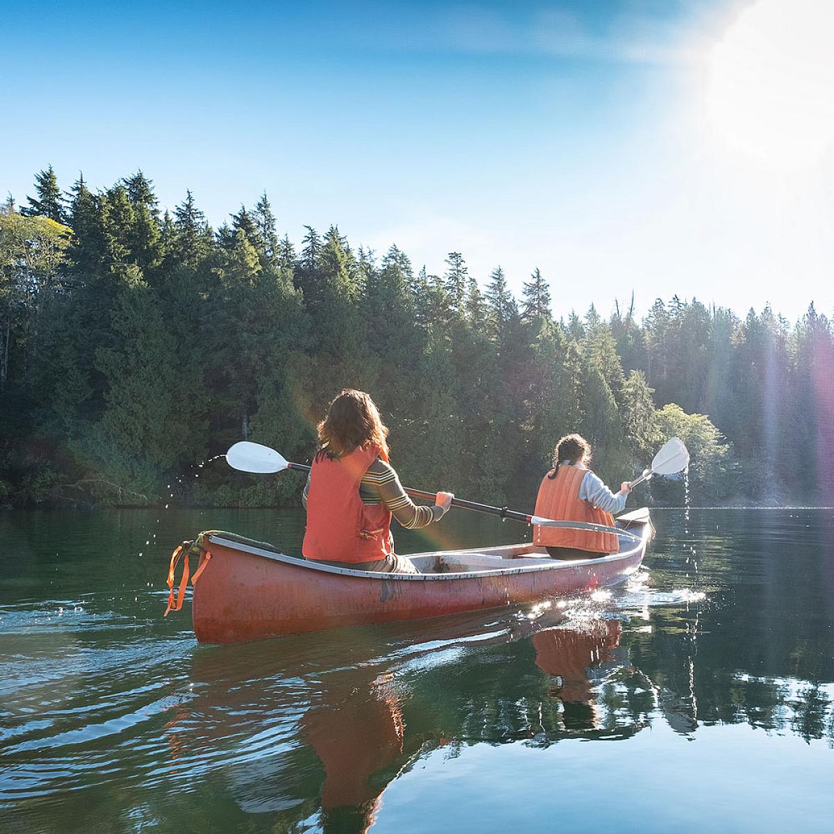 Two women paddling away from the camera in an orange canoe. They are wearing orange life vests and there are pine trees in the background.