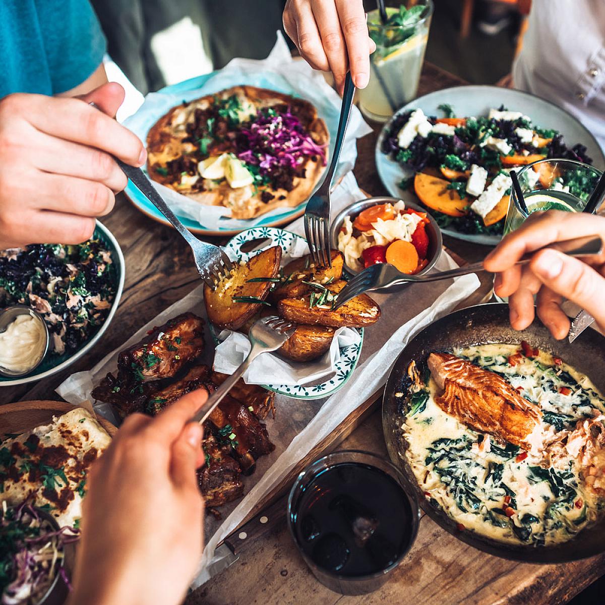 A wooden table full of food. Four separate hands reach in from the corners of the photo with utensils.