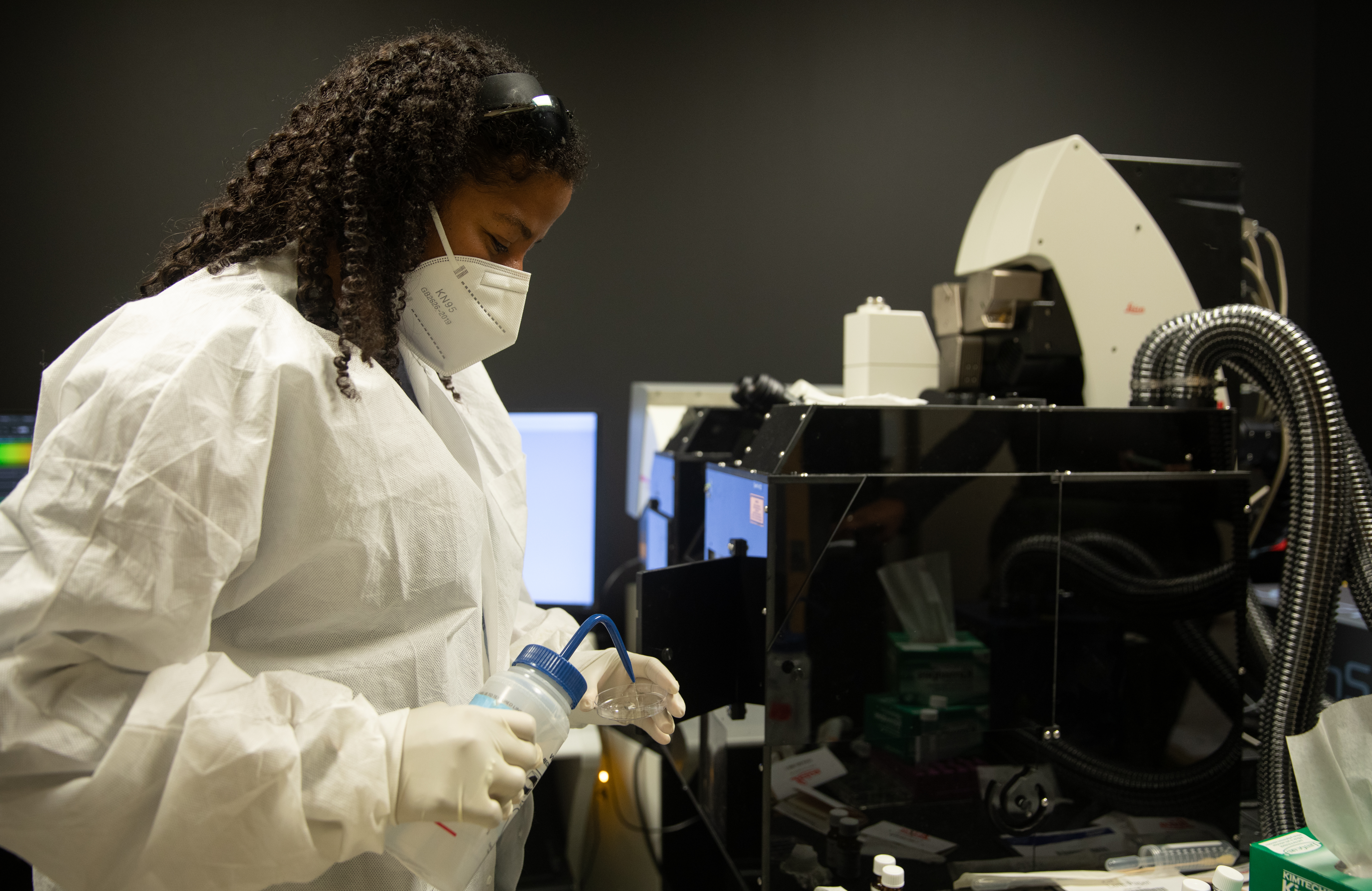 A woman with dark skin wearing a lab coat and face mask adds water to a petri dish next to a complicated-looking machine.