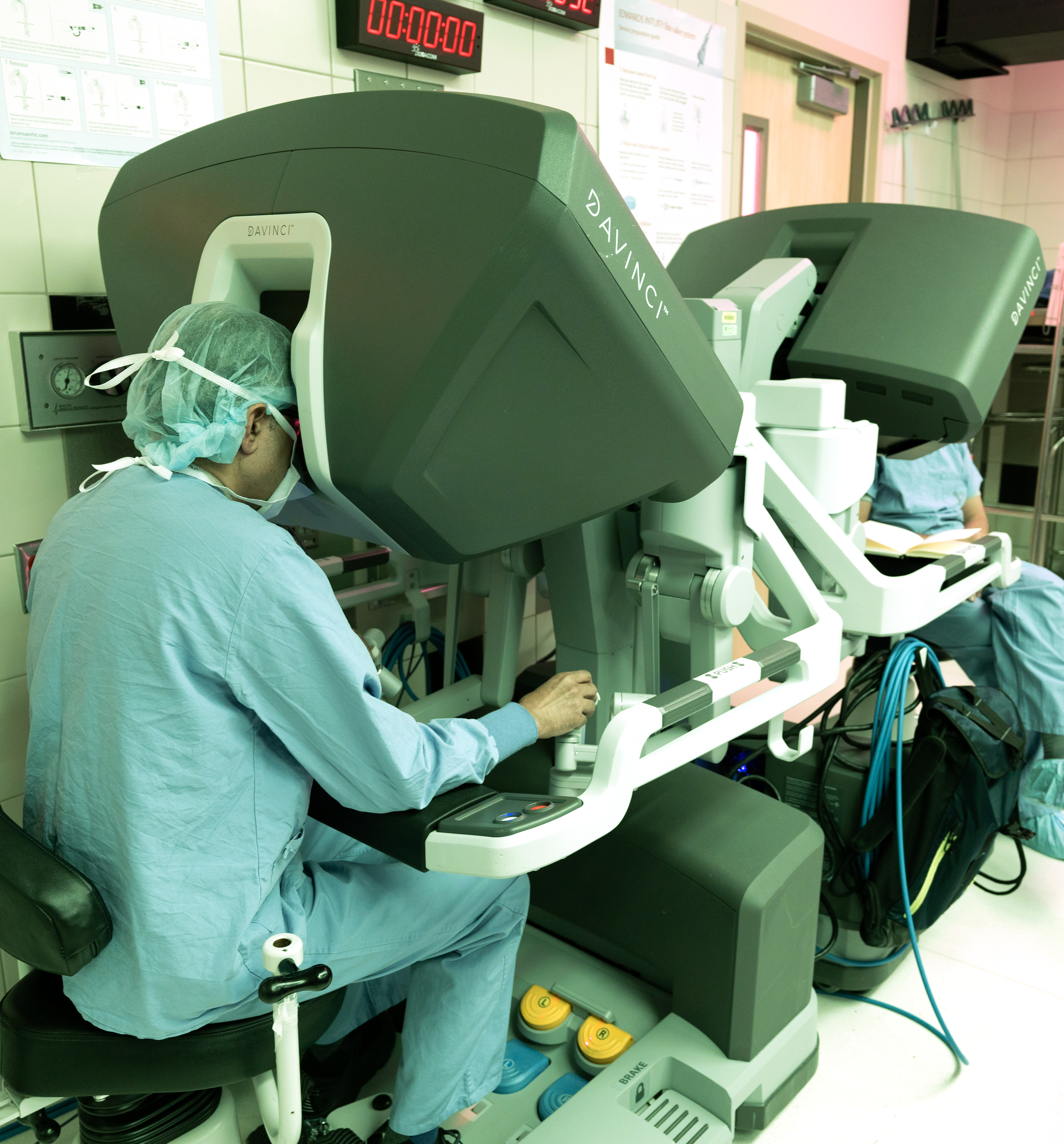 Person wearing scrubs, sitting at a controller for a surgical robot, using hand controls while peering into a viewscreen that half-envelops their head.