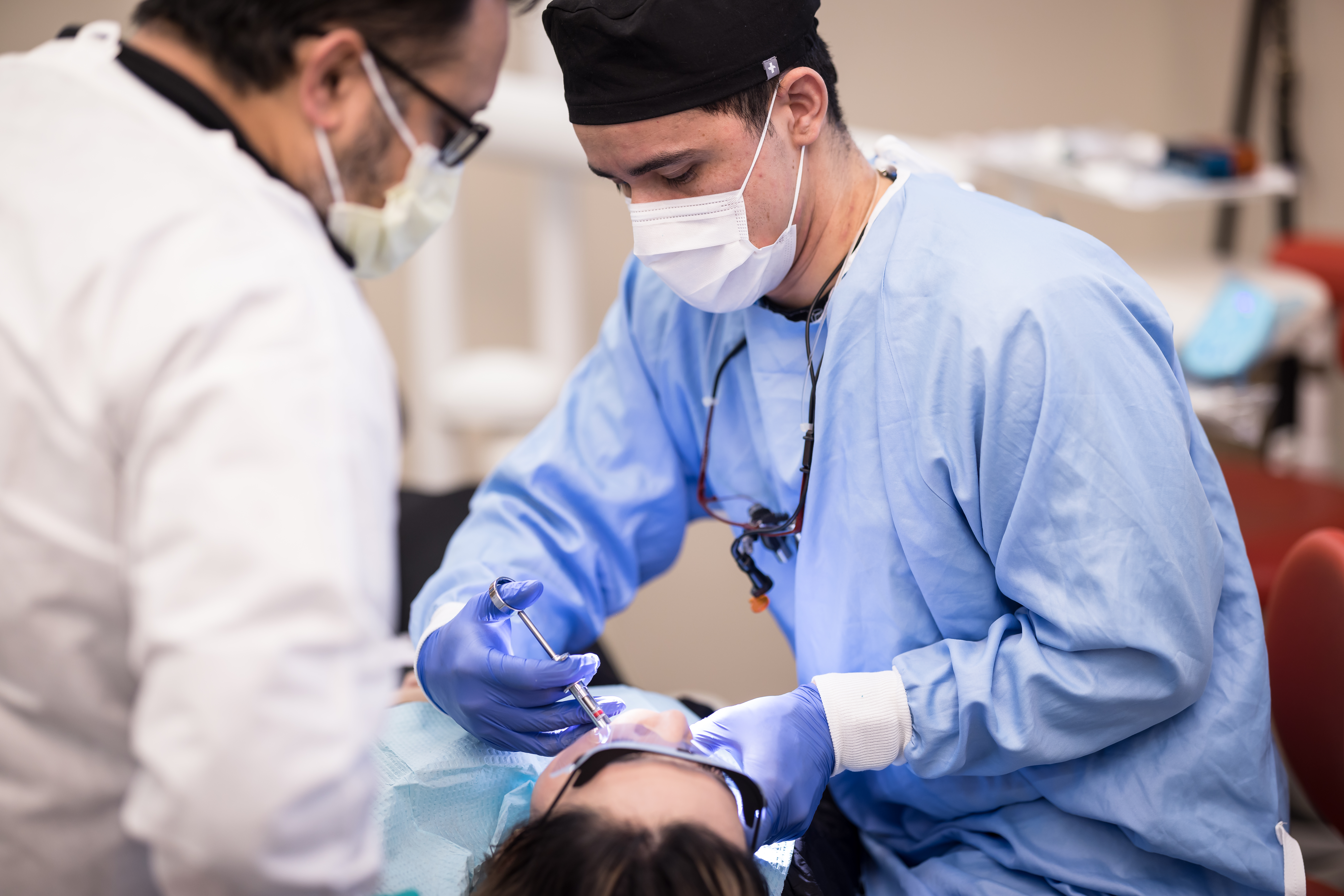 Two doctors perform a wisdom tooth extraction on a prone patient.