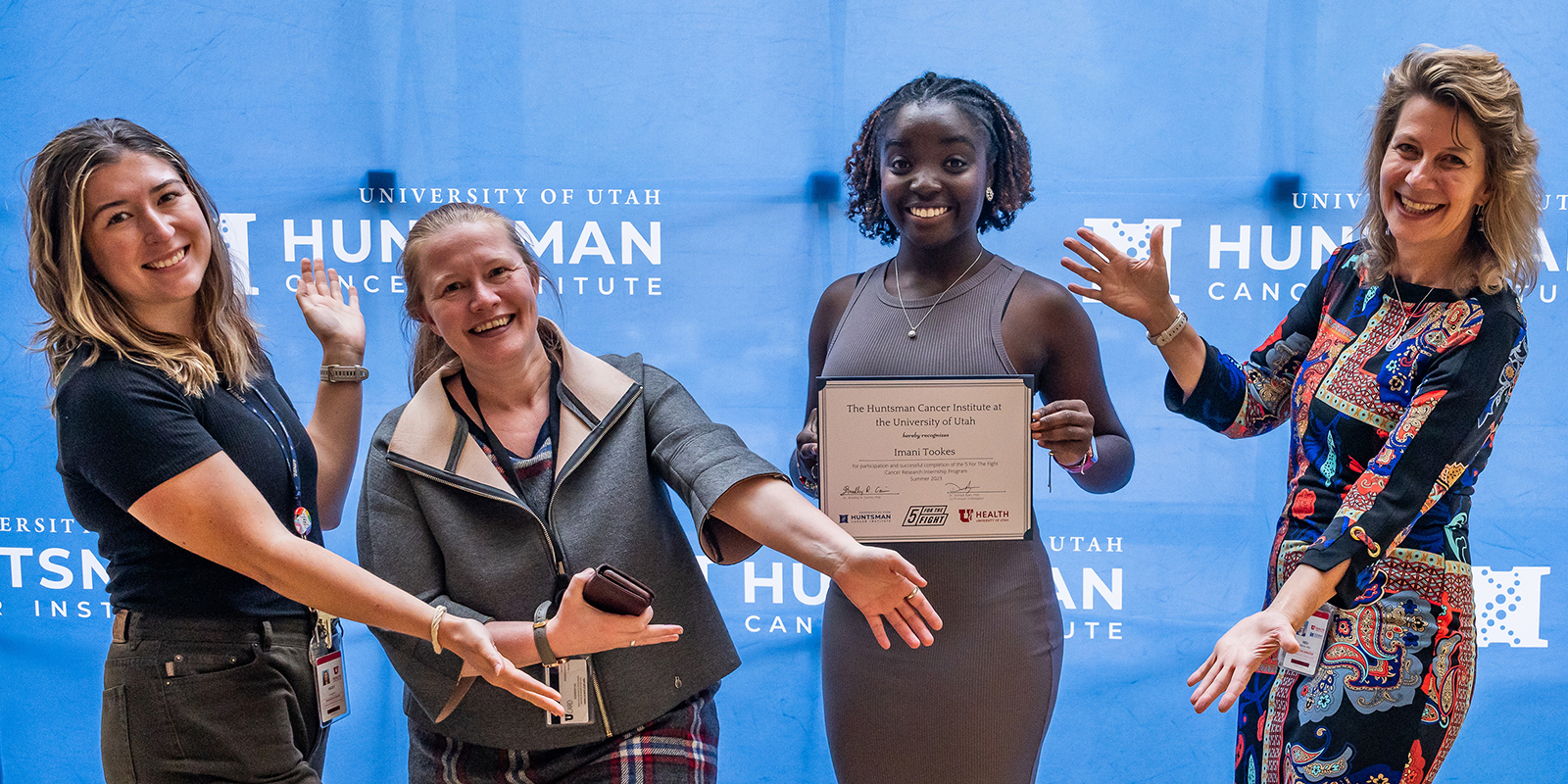 Ulrich Group members standing in front of a step and repeat backdrop and gesturing towards Imani Tookes, who is holding up a certificate of completion of the 5 For The Fight Cancer Research Internship program.