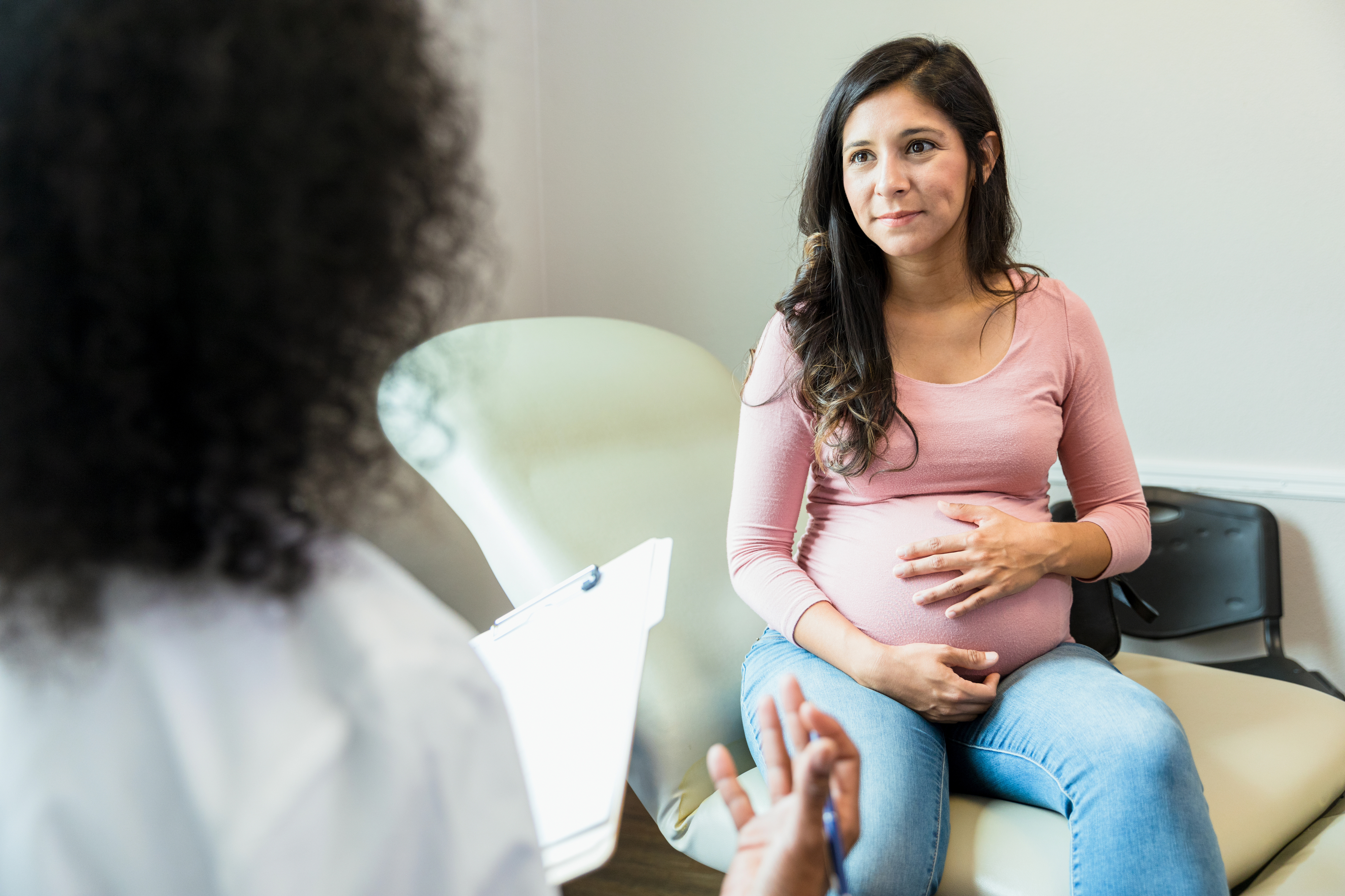 Pregnant woman with long dark hair sitting in a clinic room talking to her doctor.