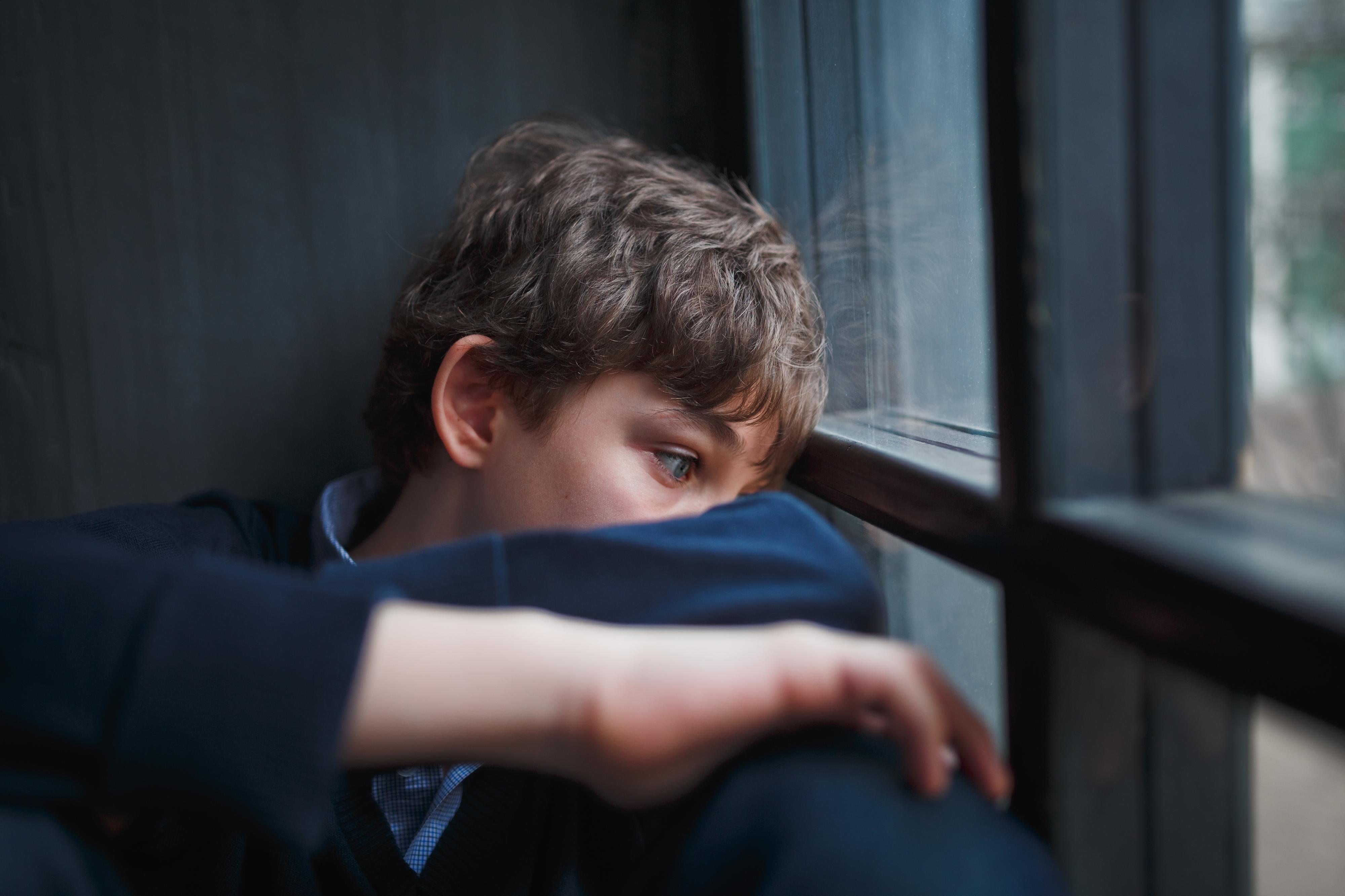 Teen boy looks depressed and is gazing out the window. He is wearing a dark blue sweatshirt and has brown hair and blue eyes.