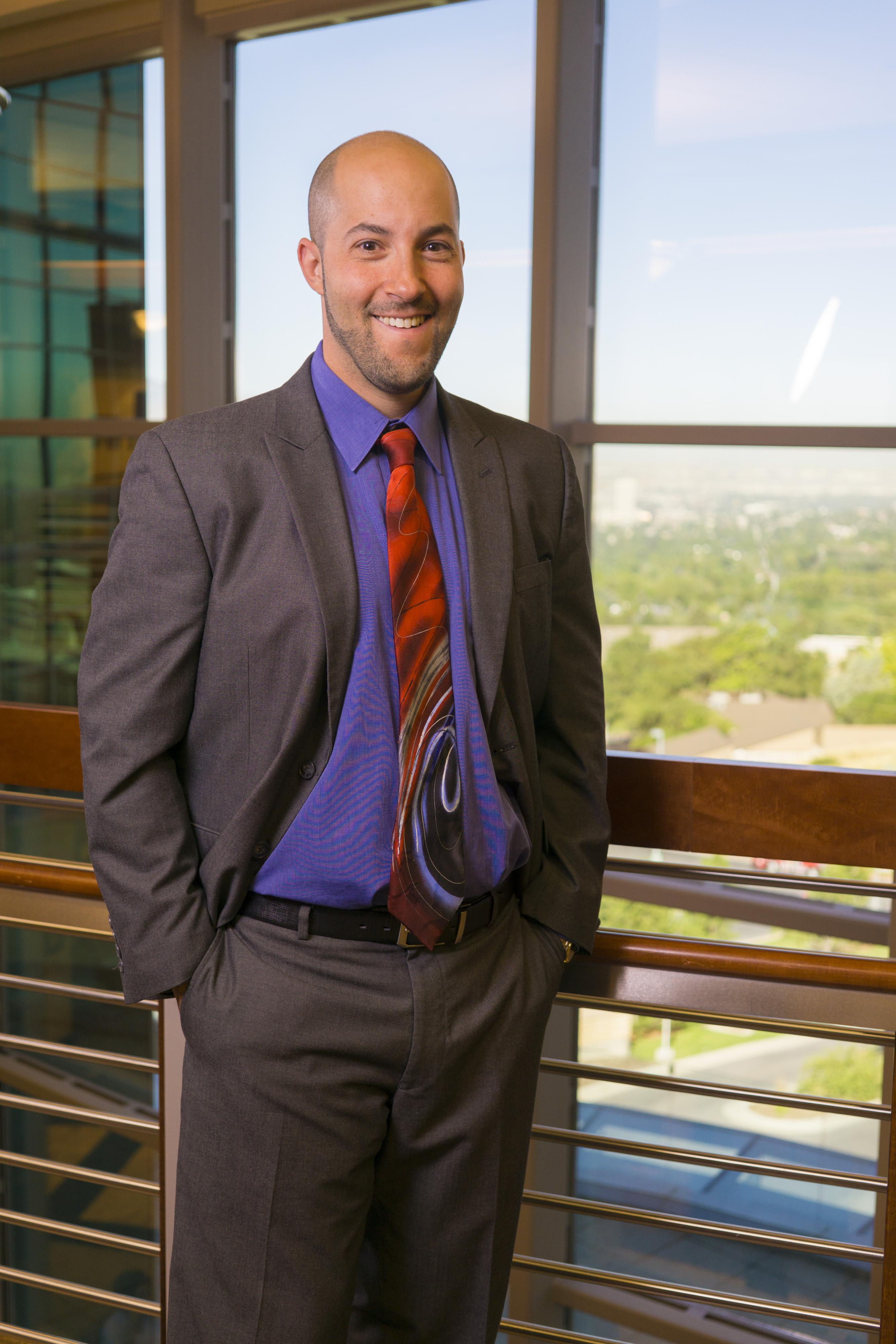 A bald person in a purple shirt and black suit grins at the camera in front of a sunny window.