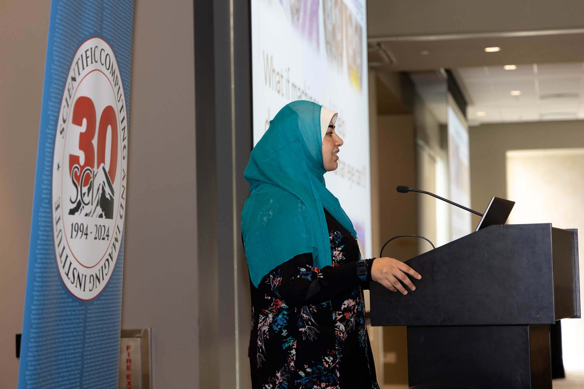 A woman in a blue hijab speaks behind a podium.