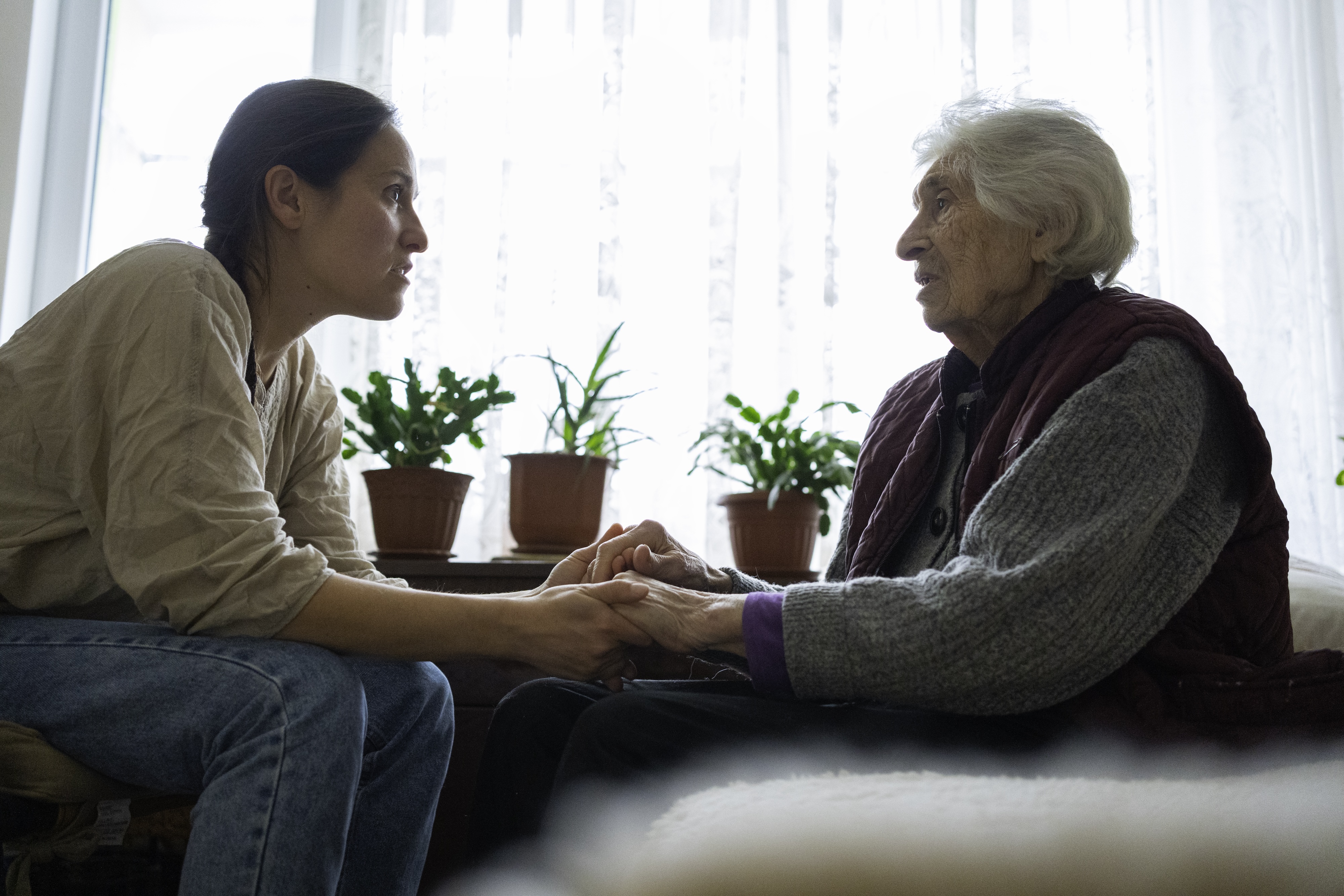 A person with brown hair and a person with white hair hold hands in front of a window, conversing intently.