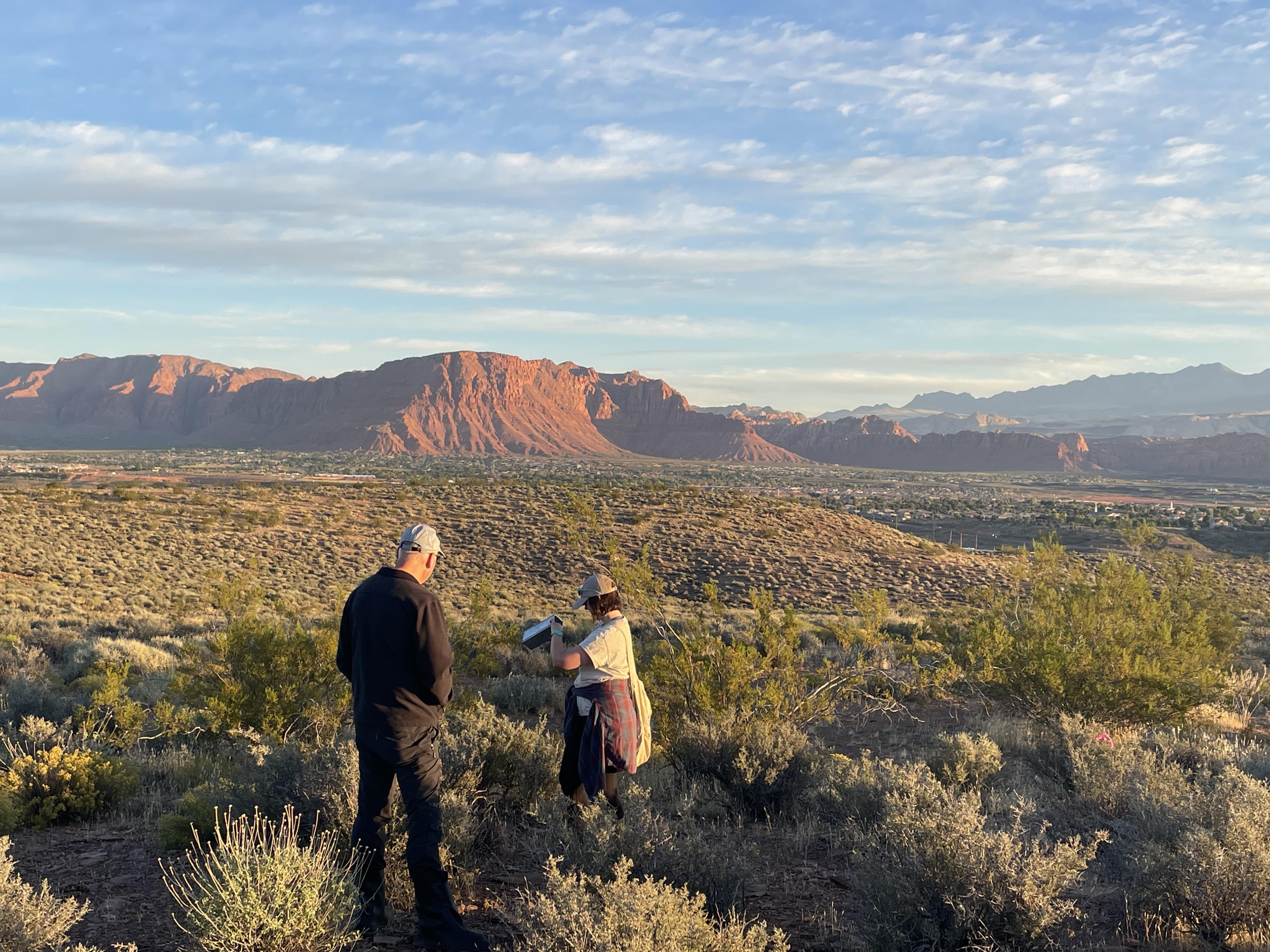 Two figures stand in a vast, scrub-covered flatland with huge mesas and mountains in the blue distance