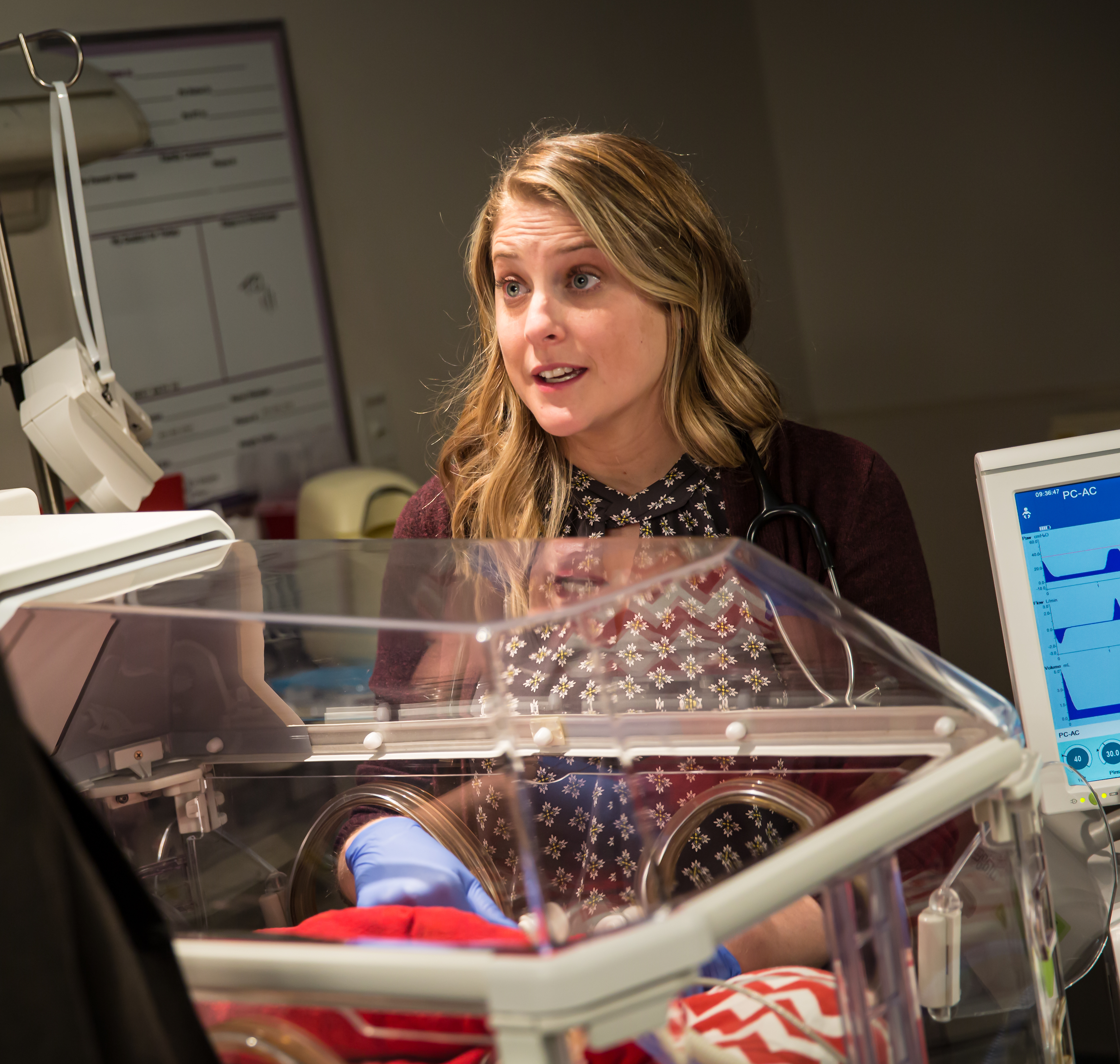 A blond woman with light skin speaks with someone out of frame in the NICU. Her hands are in an incubator. 