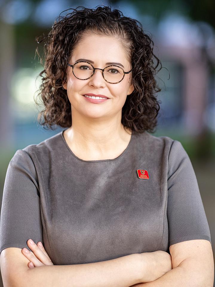 Kelly Tappenden standing with folded arms in front of her chest, she has shoulder-length curly hair and wearing round framed glasses