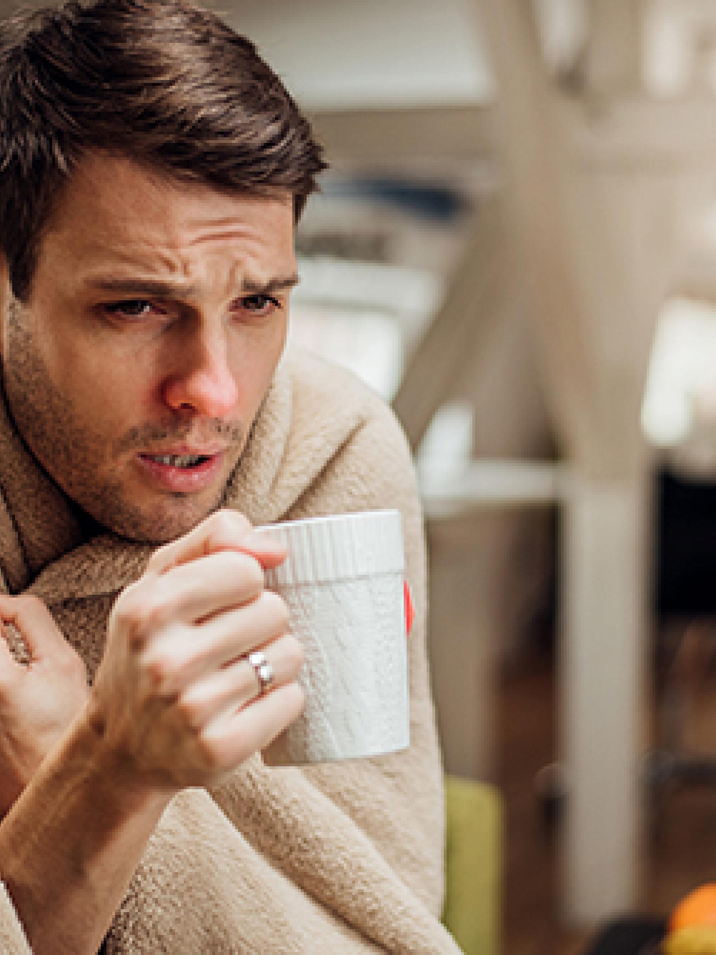 Man with a head cold holding a mug and wearing a blanket