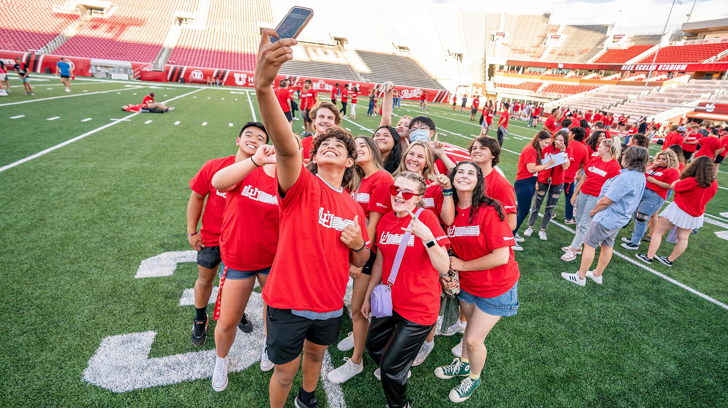 a group of freshmen in matching t-shirts take a selfie on the Rice-Eccles Stadium football field